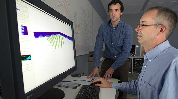 A photo of two men looking at a computer monitor that displays blue and green wind-modeling graphics.