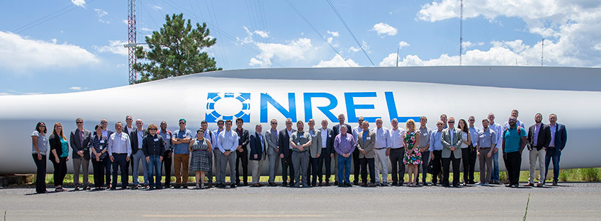 A group of people stand in front of a large wind turbine blade on the ground that has NREL's logo painted on it. 