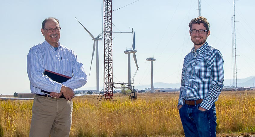 Two researchers stand in front of a series of turbines in an open field.