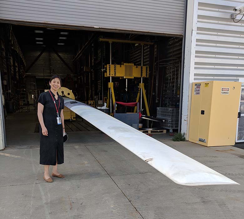 A woman stands next to a wind turbine blade jutting horizontally out of a large facility’s garage door.