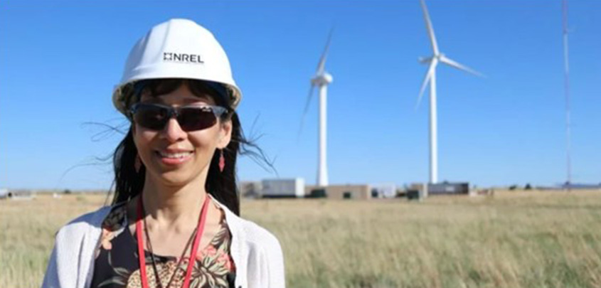 A woman stands in a field wearing a hard hat with NREL’s logo and sunglasses. Behind her are two wind turbines and several buildings. 