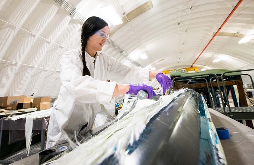 A researcher in lab coat, gloves and goggles, adjusts material on a wind blade in a hangar