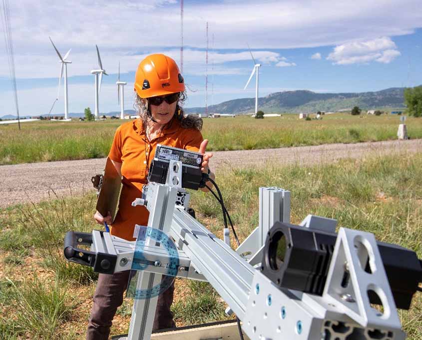 A woman in a hard hat and sunglasses operates equipment on a grassy plain in front  of wind turbines and mountains