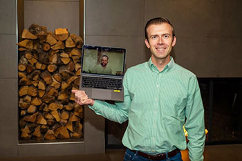Jared Thomas stands in front of a fireplace and a stack of logs while holding up a laptop showing Christopher Bay