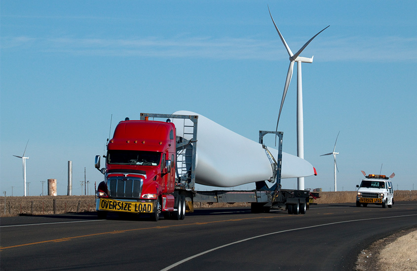 A truck drives a wind turbine blade past a wind turbine.