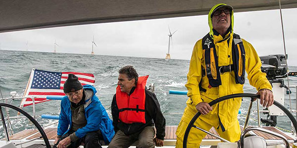 Three people on a boat, one driving, in front of offshore wind turbines.