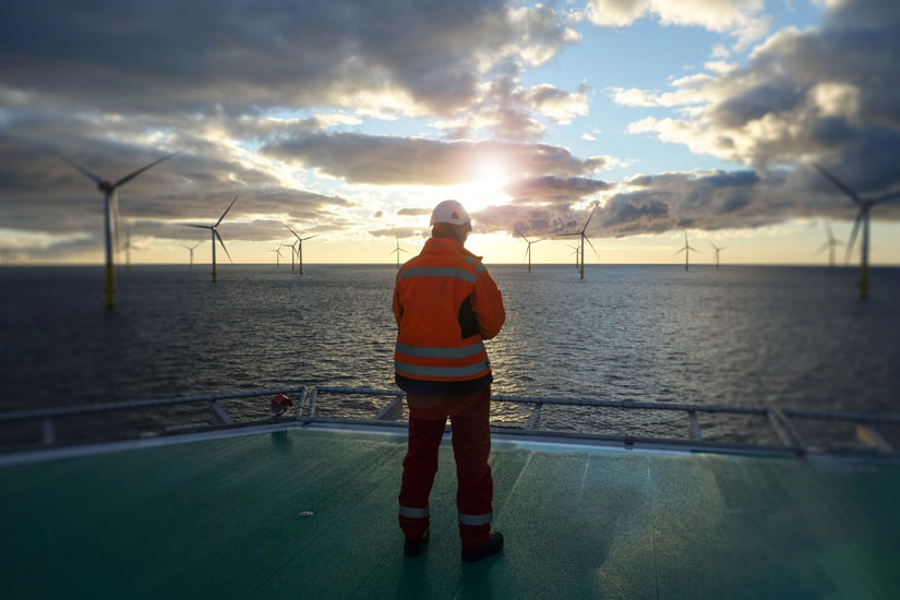 Offshore manual worker standing on helipad with wind-turbines behind him in sunset.