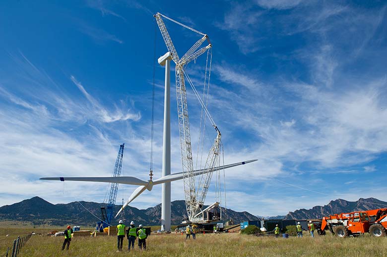 A crane lifts three connected wind turbine blades next to a tower as several workers look on