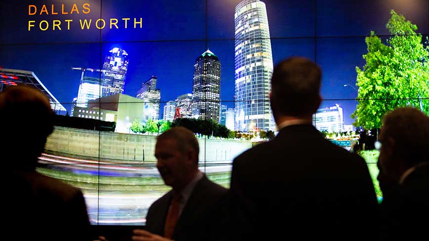 Silhouettes of several people walking in front of a large monitor with an image of a cityscape and text that reads, "Dallas Fort Worth."