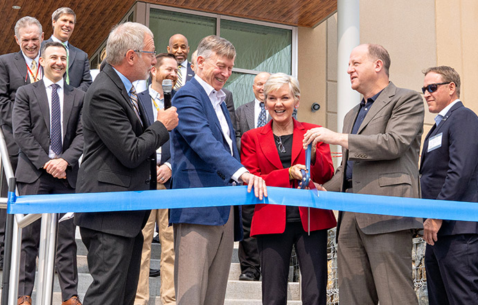 Photo of a group of people standing behind a ceremonial blue ribbon in front of a new building while two people cut the ribbon with an oversized pair of scissors