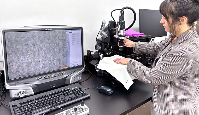 A person examines a fabric under a microscope next to a computer screen.