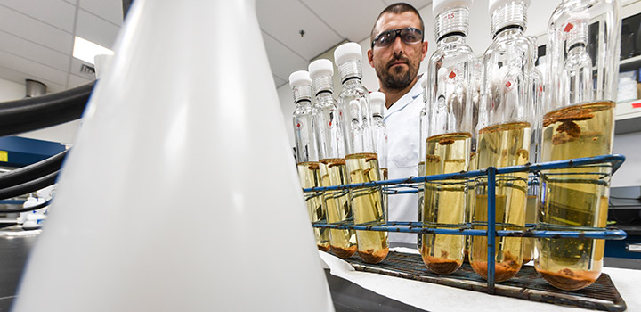 A man in safety glasses and lab coat stands behind a row of glass vials with yellow liquid in a laboratory.