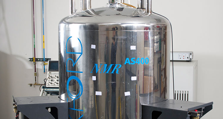 Photo of a 400 MHz Agilent Unity Inova NMR Spectrometer, which is composed of a large metal tank on a three-legged metal stand; clear plastic tubes and wires stick out of the top and bottom of the tank.