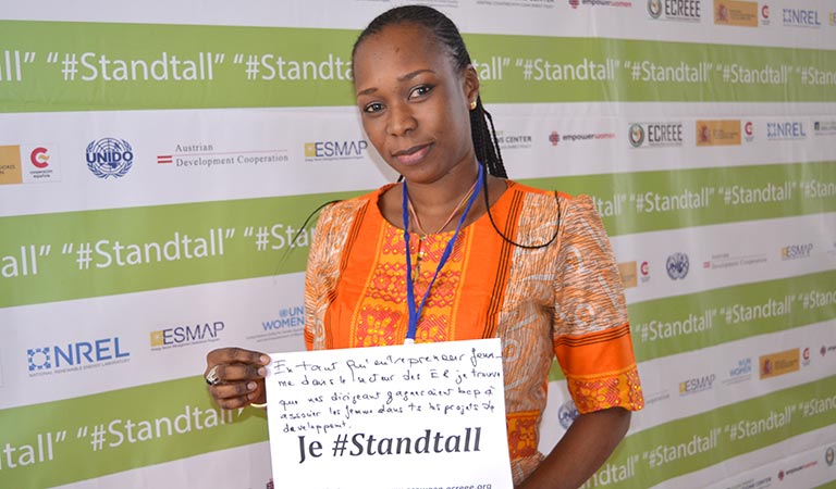 Photo of a black woman in an orange dress holding a piece of white paper that says: "Je #Standtall"