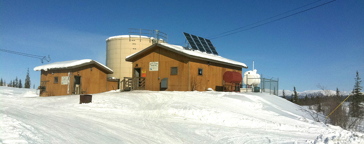 A washeteria in Alaska with solar panels on the roof and a propane tank and water tank next to the building.