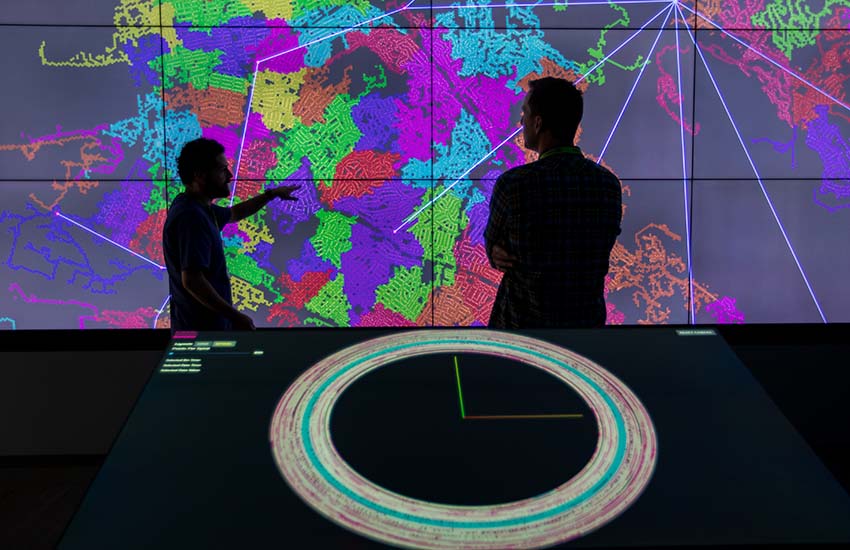 Two people pointing and looking at dataset on large computer screen.