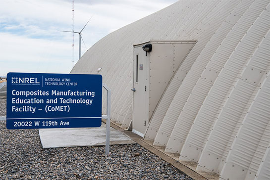 Photo of a white dome-shaped building with a sign that says, 'Composites Manufacturing Education and Technology Facility – (CoMET ),' and a wind turbine in the background. 
