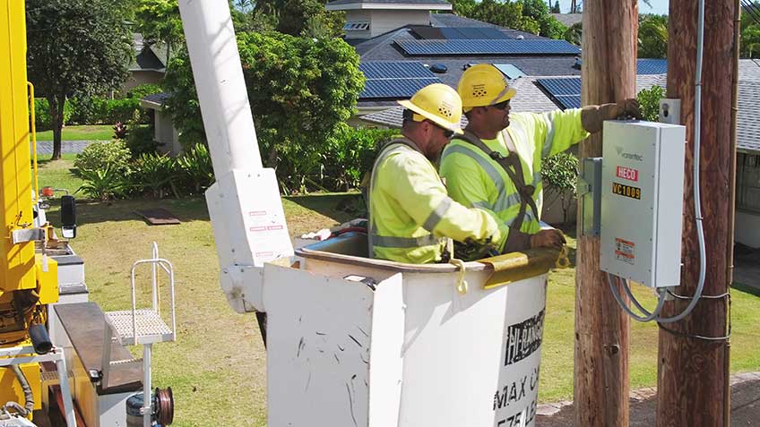 Photo of two men in yellow hard hats installing electrical equipment on a telephone pole.