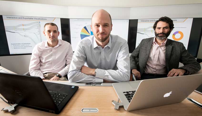 Three men sit at a desk with two laptops in front of them.